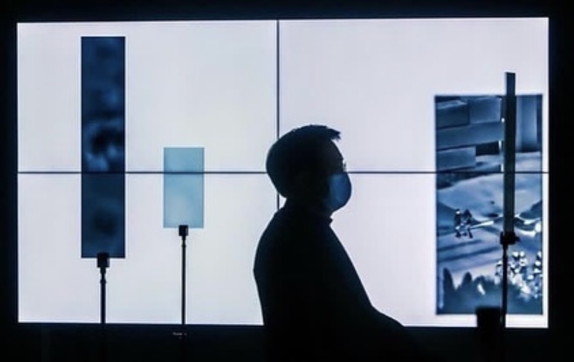silhouette of a person in front of white tv screens with smaller screens in front revealing parts of an image on the white screens