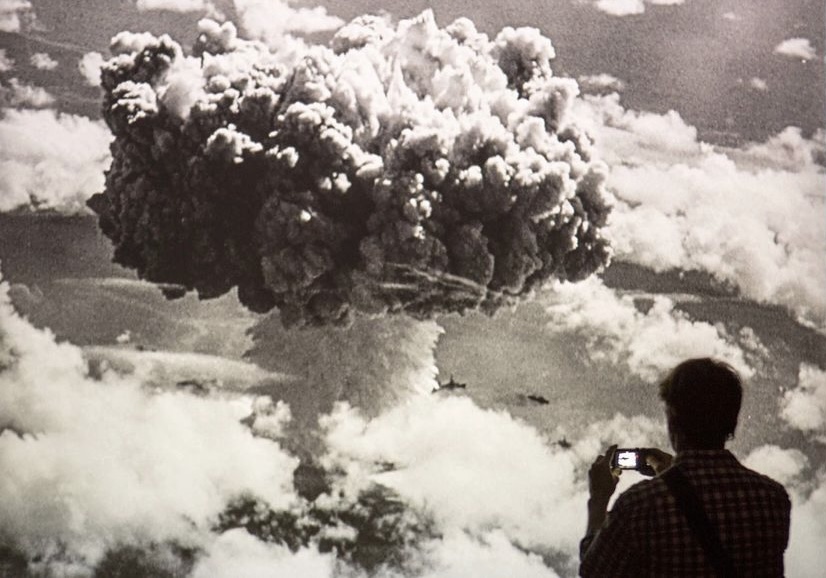 a silhouette of a man taking a picture with a phone in front of a black and white oversized image of an atomic bomb explosion