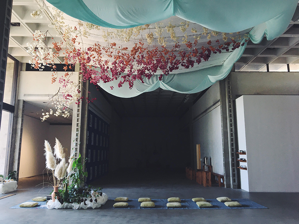 A large room with pillows lined up on the floor and hanging textiles and flowers above