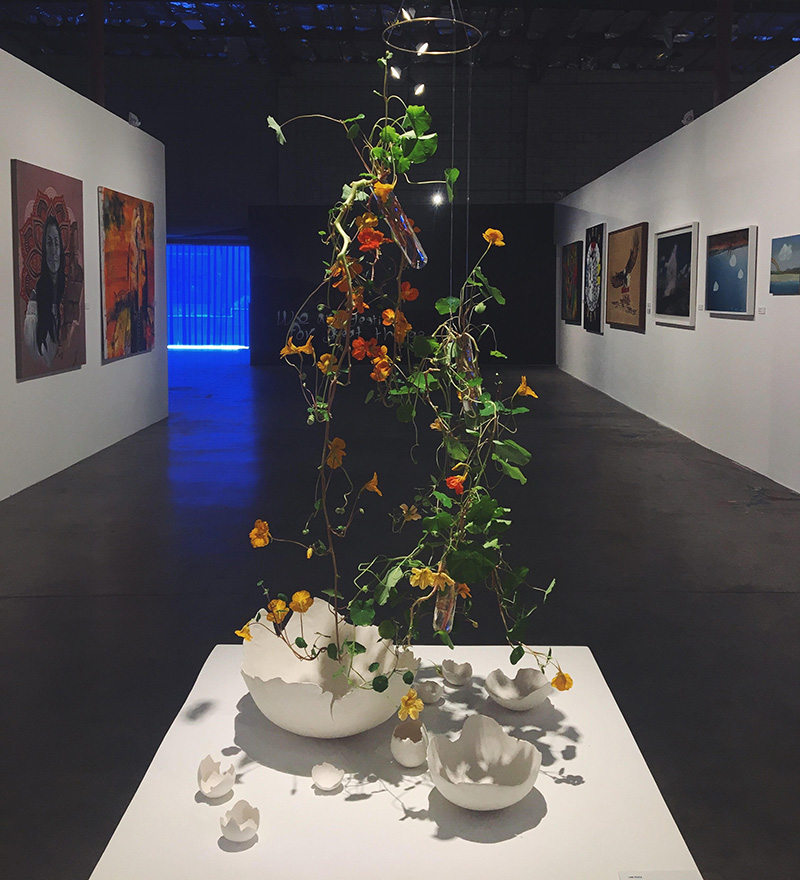Dark gallery space with a hanging mobile of flowers hanging above a large broken egg shell