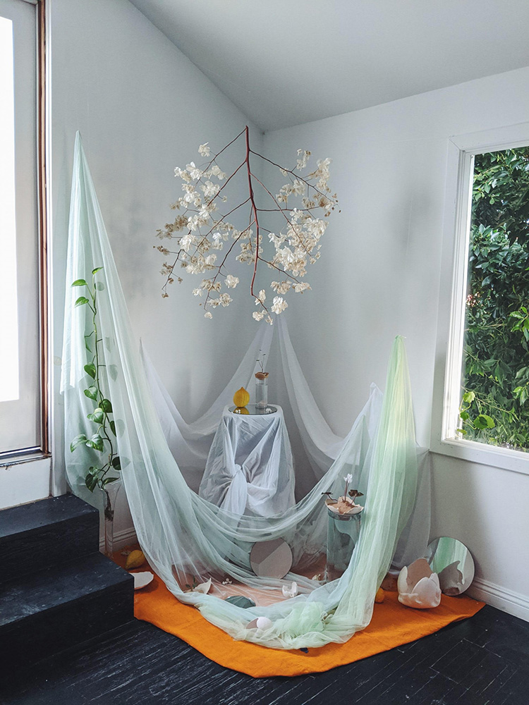 the corner of a bright room with a hanging branch and white flowers above draped white and green fabric and an orange rug